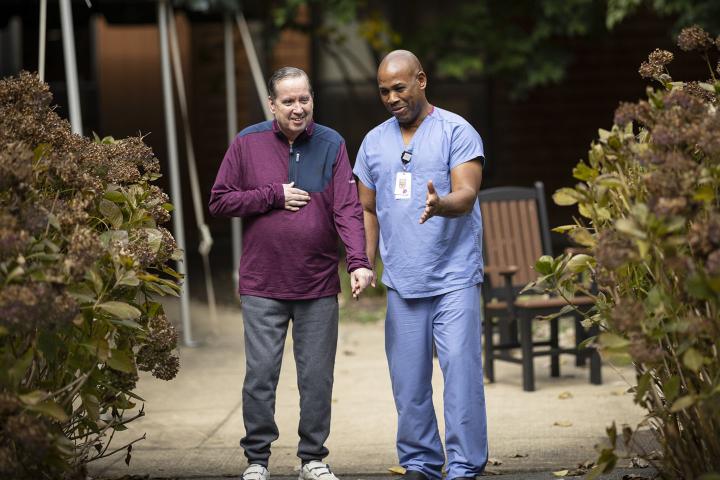 A male certified nurse assistant walks with a patient in an outdoor patio.