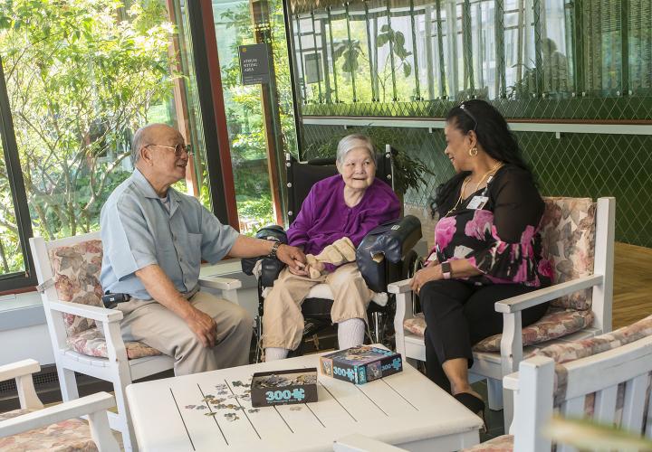 An older couple sit with a visitor in a bright sitting area.