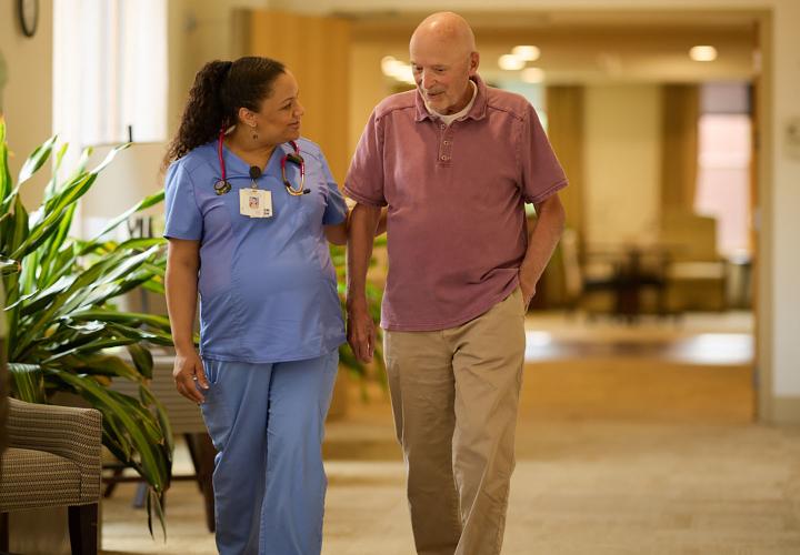 A female nurse assists an older male patient walking down a hallway.