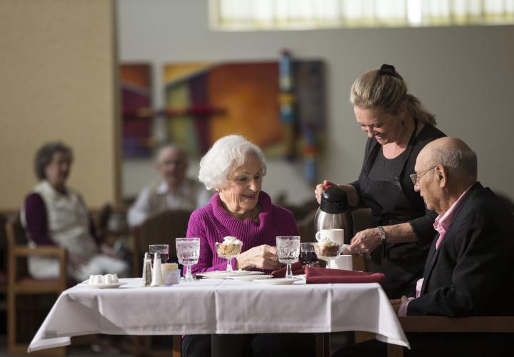 Two older residents talk with a waitperson while enjoying dinner.