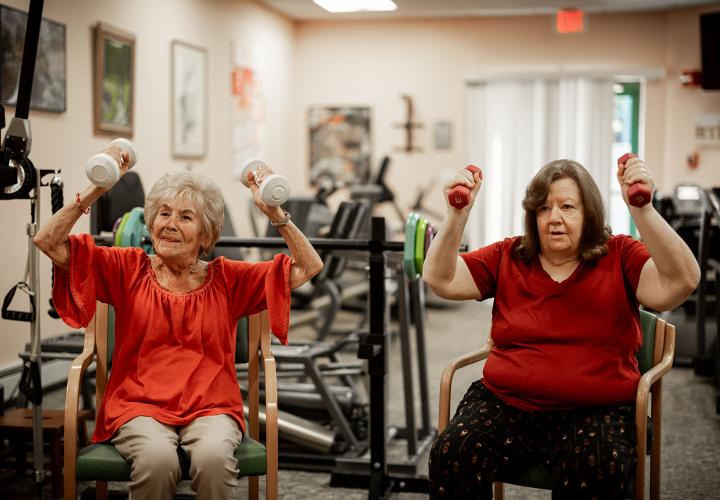 Two older women lift hand weights in a fitness room.