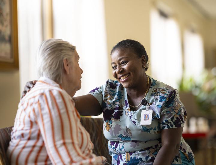 A patient care assistant smiles while speaking with an older female patient.