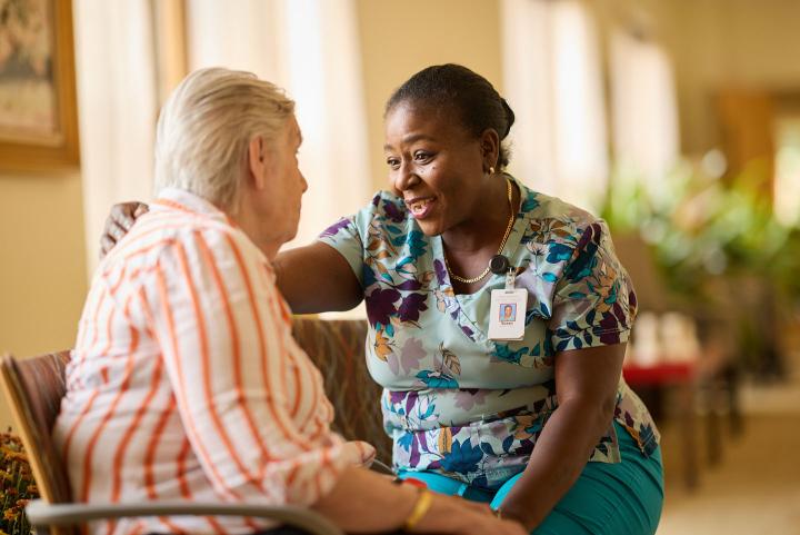 A patient care assistant smiles while speaking with an older female patient.