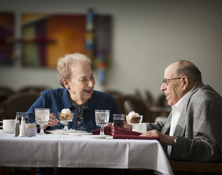 An older man and woman are seated at a restaurant table, smiling and looking at each other. There is dessert in front of them and the woman is holding a spoon.