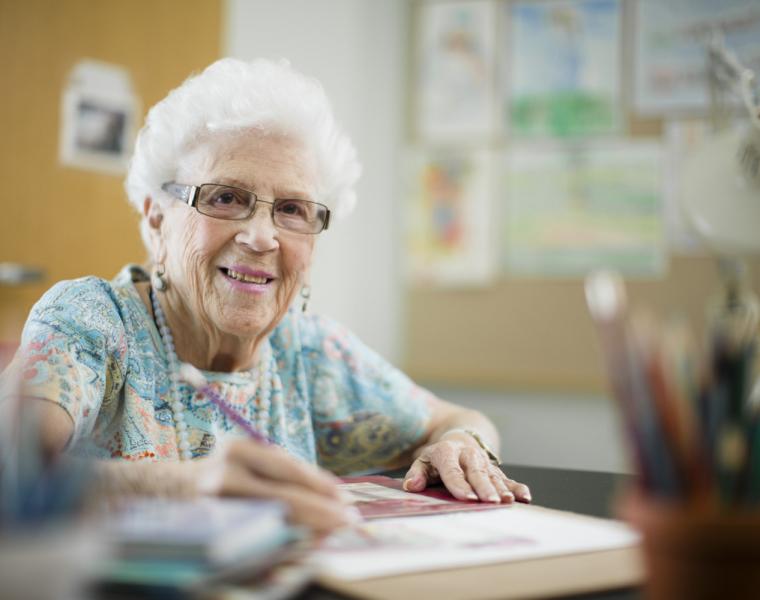 An older woman sits at a table, holding a pencil, sketching on a notepad. There is a jar of colored pencils in the foreground and artwork hanging on the wall behind her.