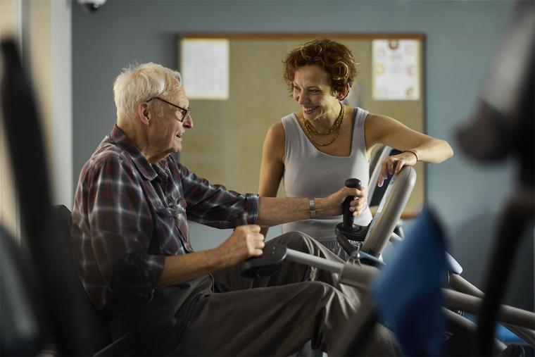 An older resident receives encouragement from a fitness expert while on an exercise bike.