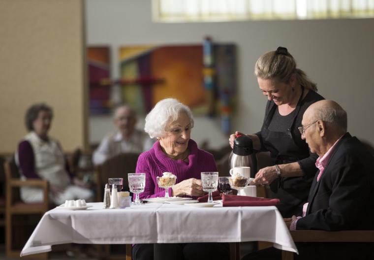 Two older residents talk with a waitperson while enjoying dinner.